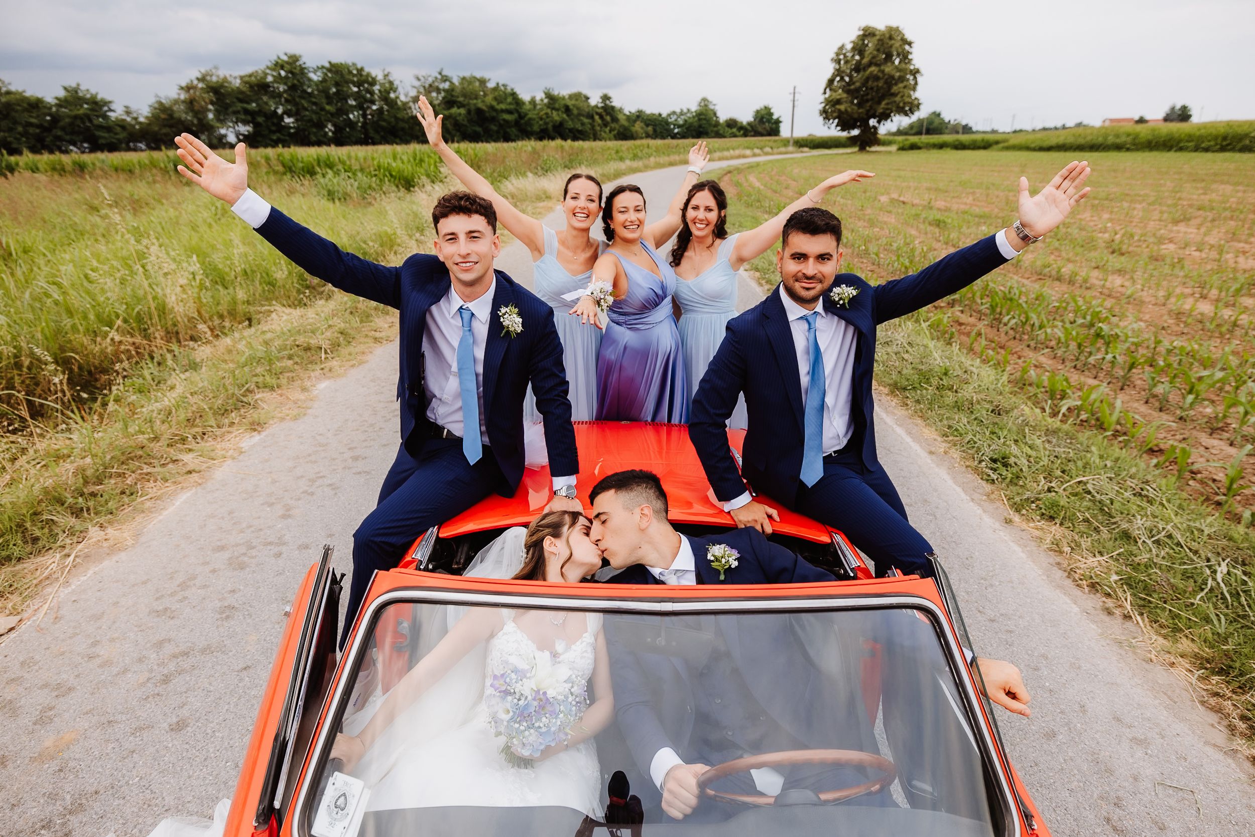 Wedding party celebrating and waving while riding in a bright orange vintage car on a country road.