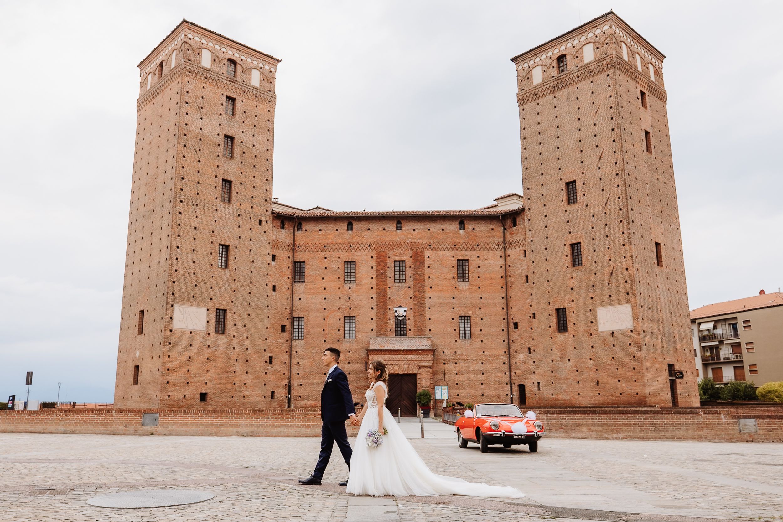 A couple stands in front of a grand historic brick castle with twin towers and a vintage red car in Bologna, Italy.