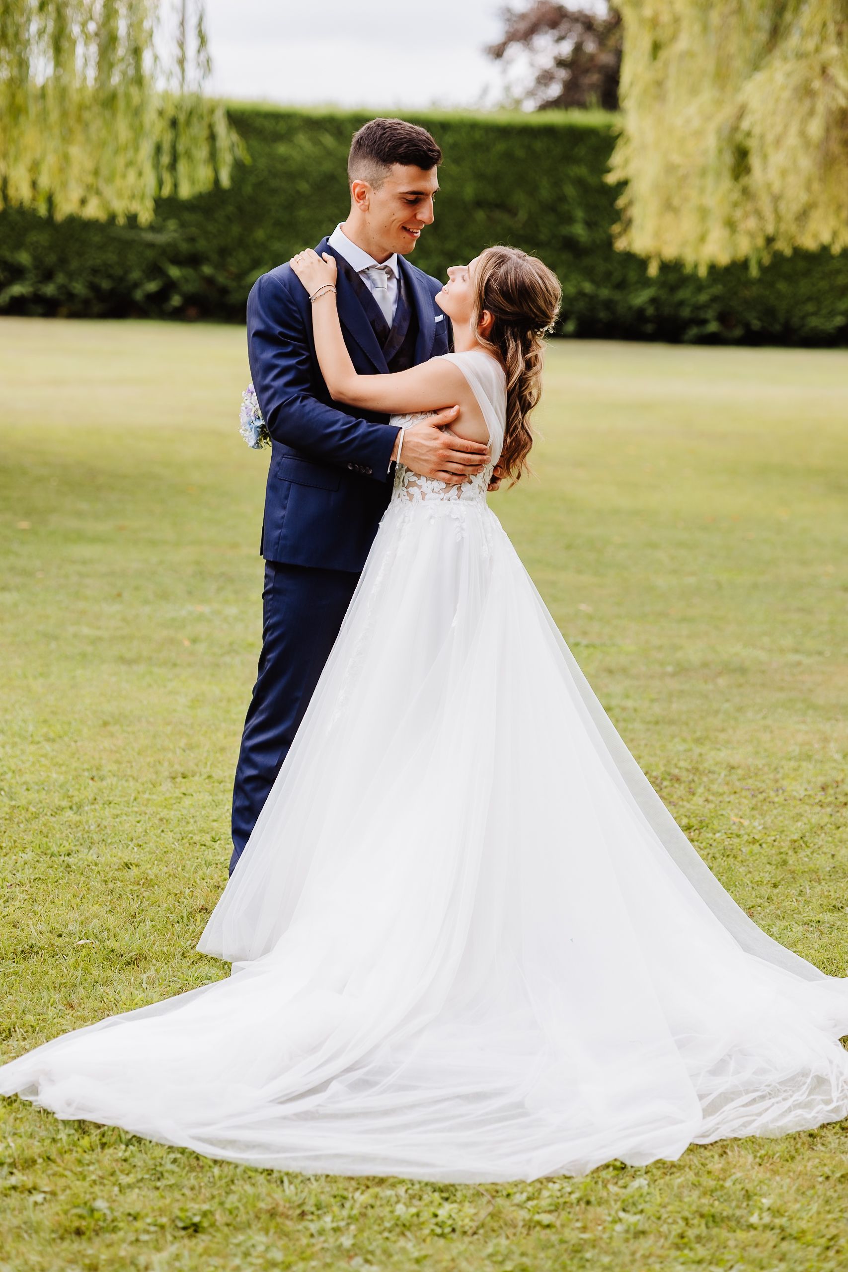 A bride and groom share an intimate moment dancing on a lush green lawn framed by drooping willow trees.