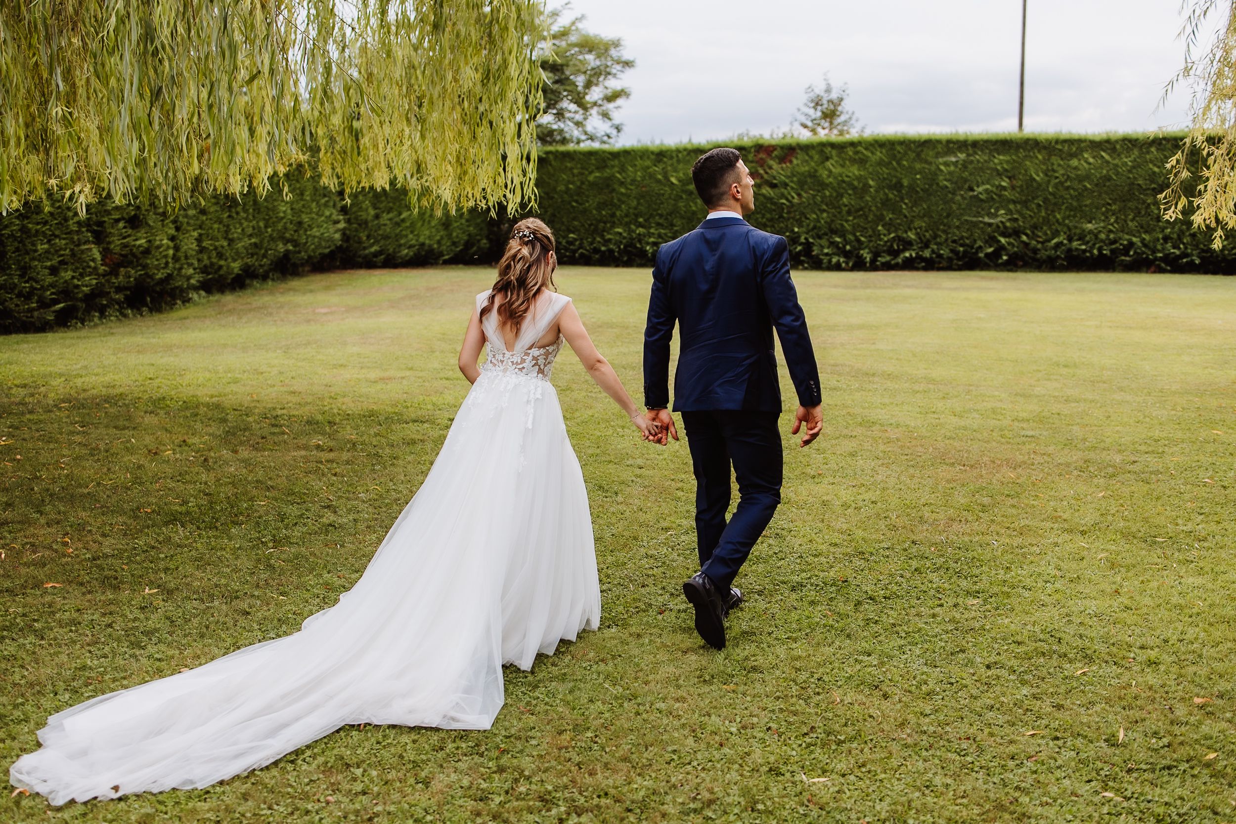 A wedding couple walks on a manicured lawn with weeping willow trees and hedges in the background.