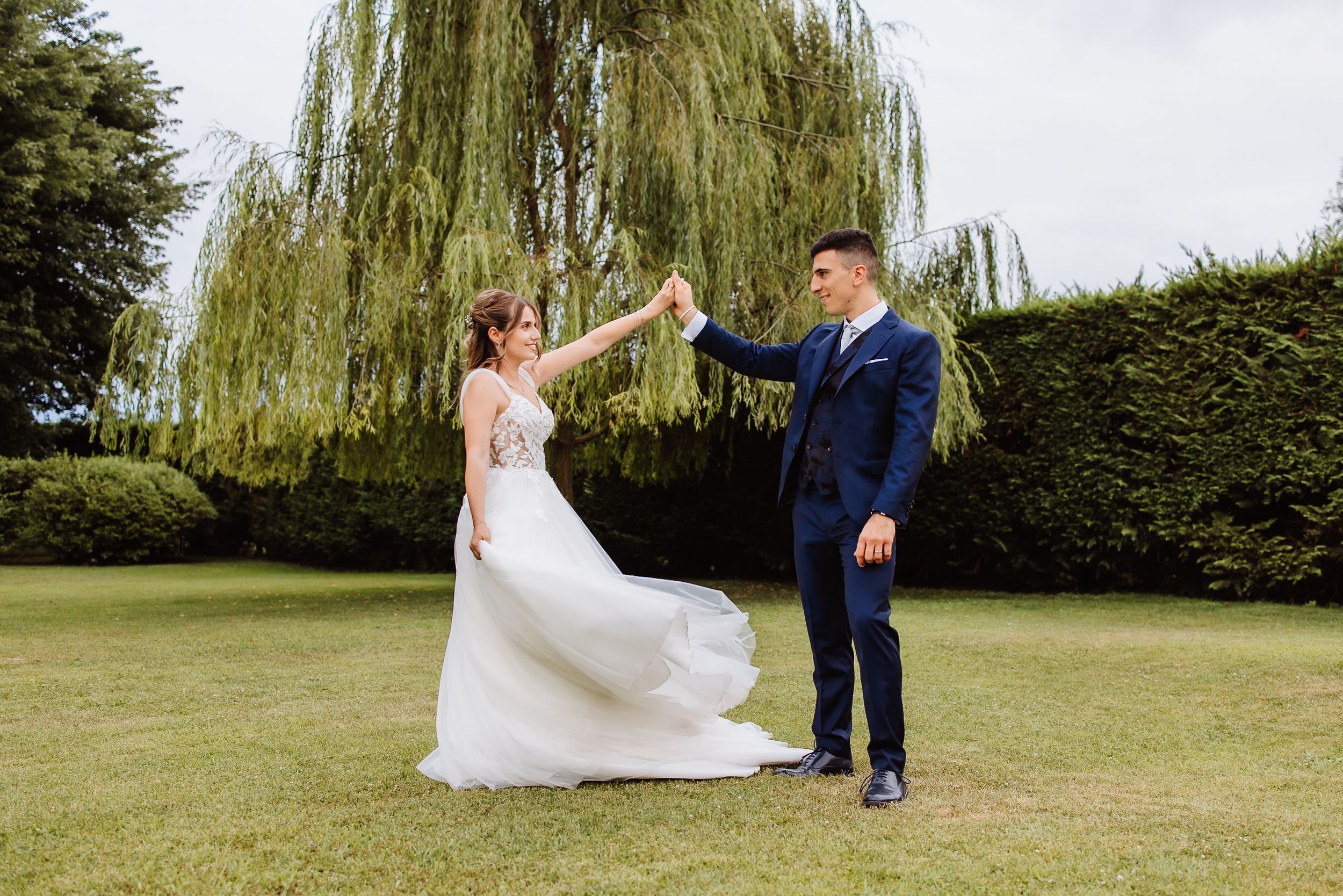 A joyful couple dancing together on a lawn with a weeping willow tree and garden setting.