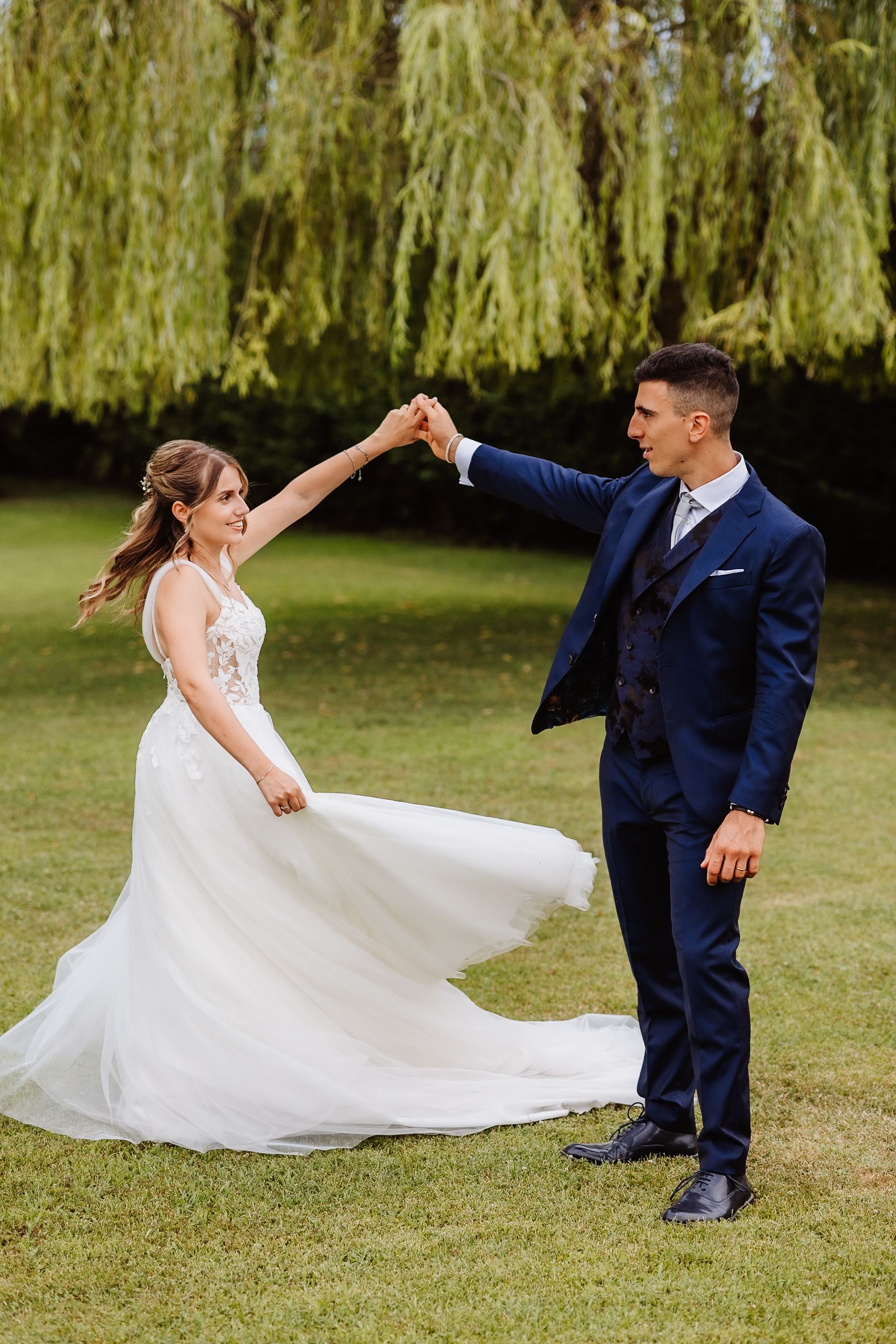 A couple spins and dances together on a lawn beneath hanging willow trees in elegant wedding attire.