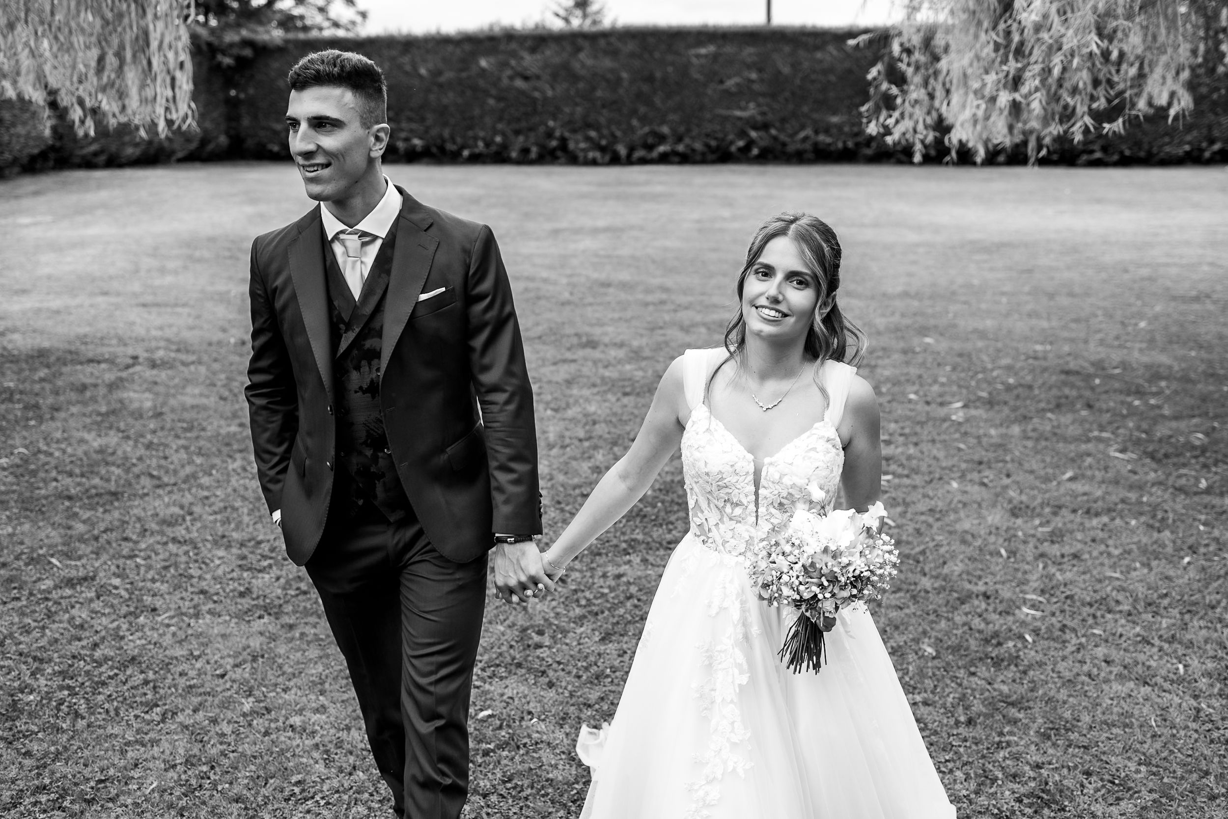 A black and white photograph of a bride and groom walking together on a manicured lawn.