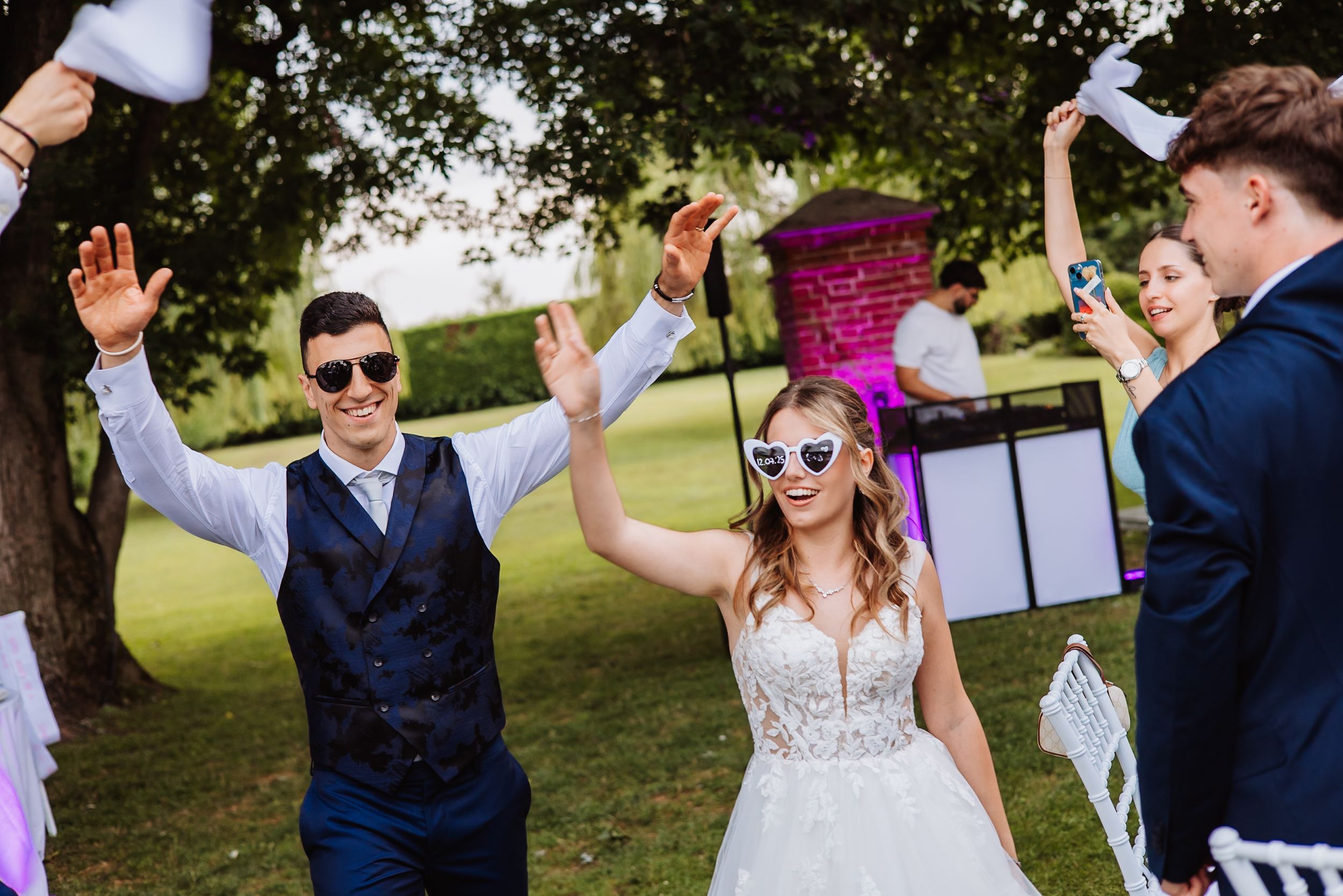 Wedding party with bridesmaids and groomsmen posing and high-fiving in a sunny outdoor garden setting.