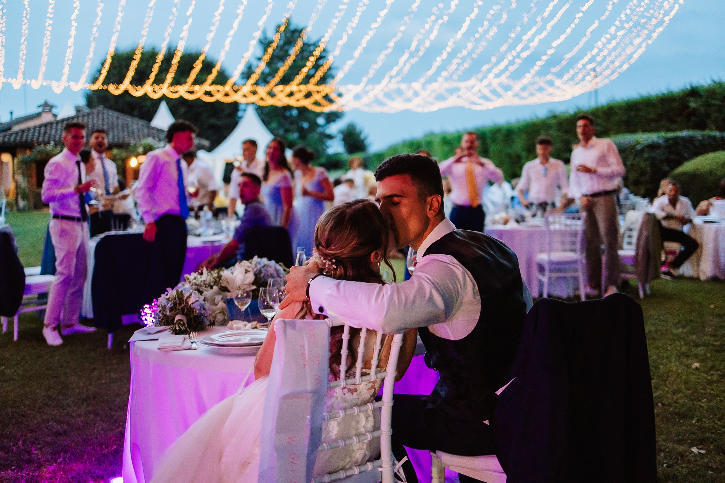 Outdoor wedding reception at dusk with string lights overhead and purple uplighting illuminating white tables and chairs.