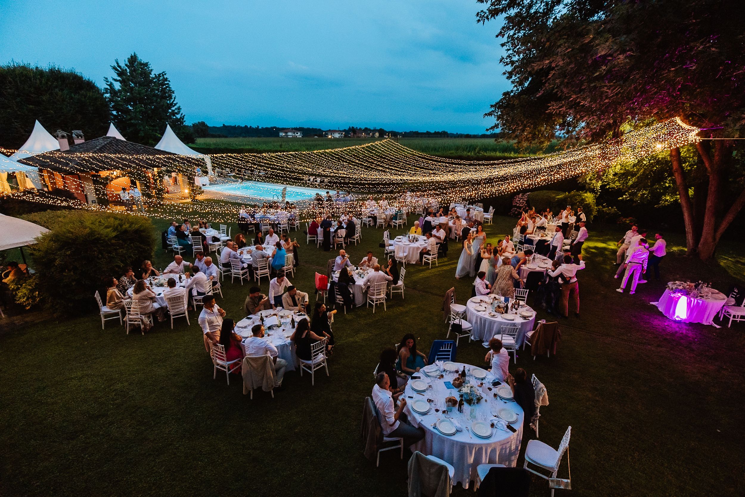 Outdoor evening wedding reception with round tables and string lights under a tent overlooking vineyard landscape.