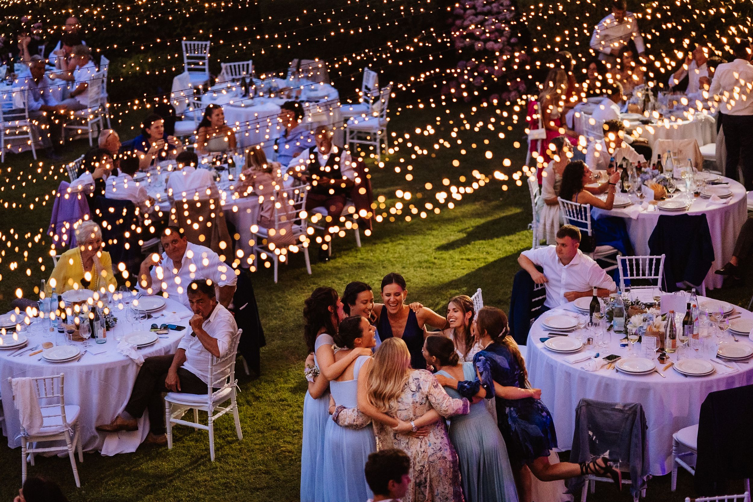 Elegant outdoor evening wedding reception with fairy lights and round tables set up on a lawn under twinkling string lights.