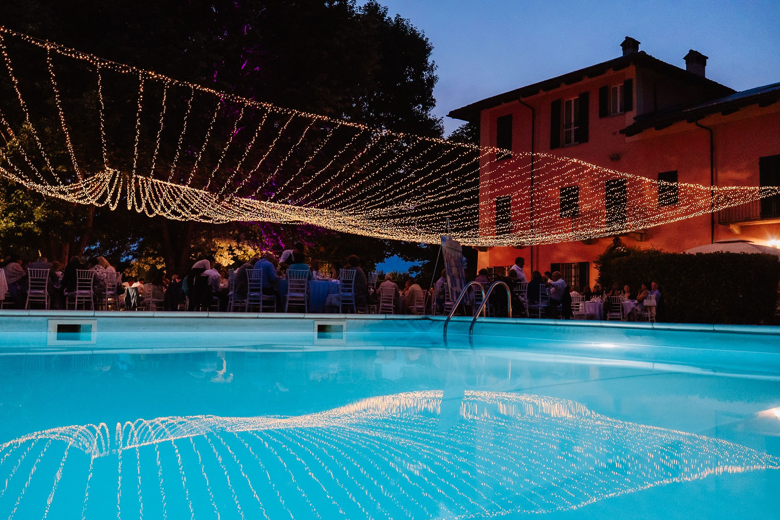 String lights illuminate an outdoor swimming pool at dusk next to a red brick building with a festive party setting.