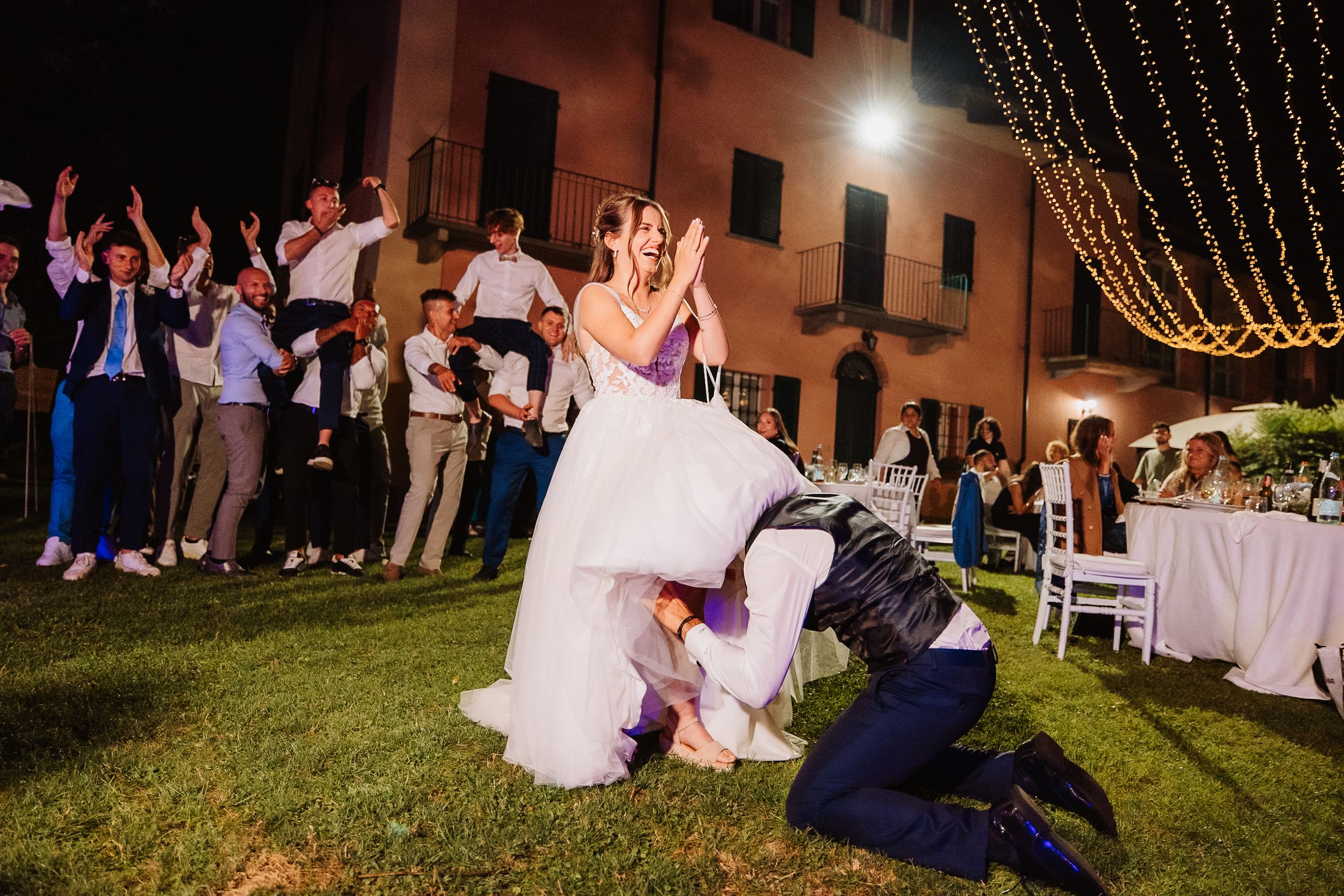 String lights illuminate an outdoor evening wedding reception with guests mingling around white-clothed tables in a courtyard.