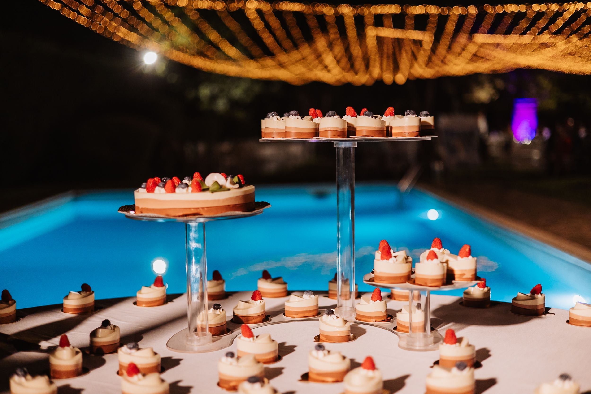 Wedding desserts displayed on tiered stands beside illuminated swimming pool with glowing orange lights overhead.