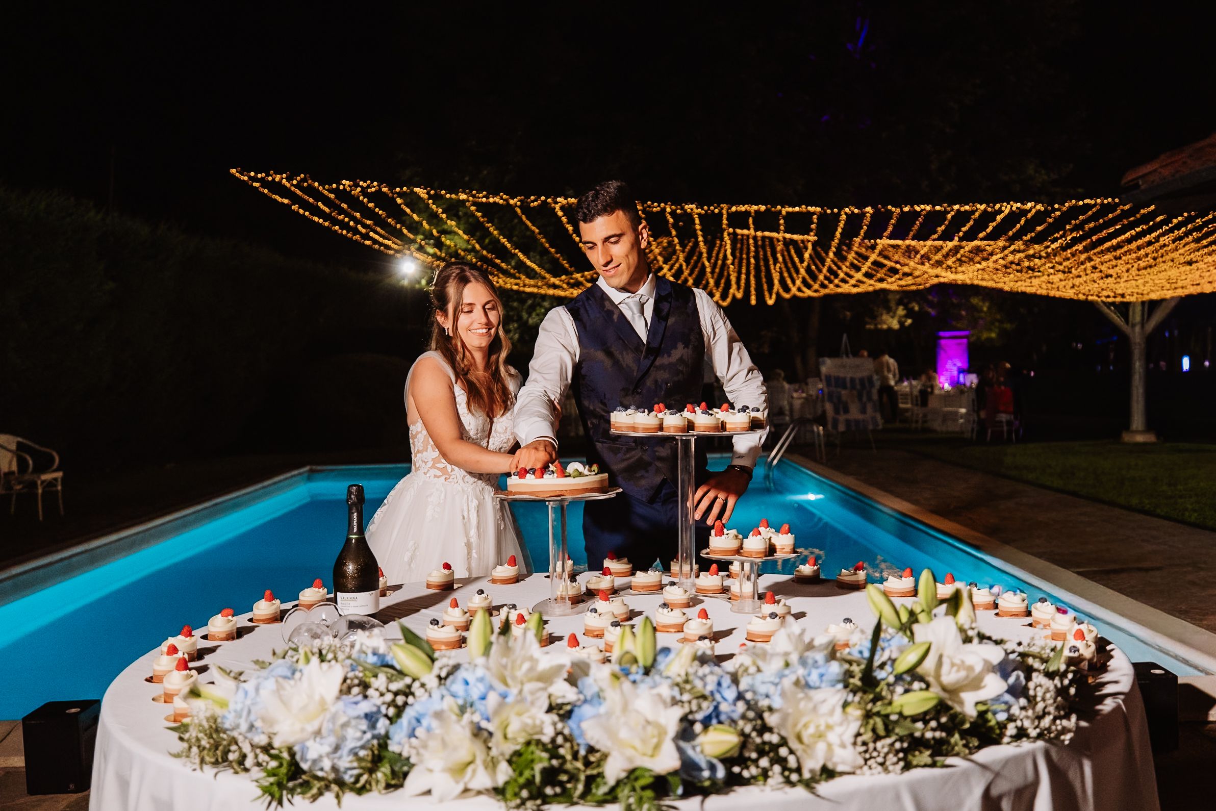 A long wedding reception table setup outdoors at night with glowing lights and floral centerpieces.