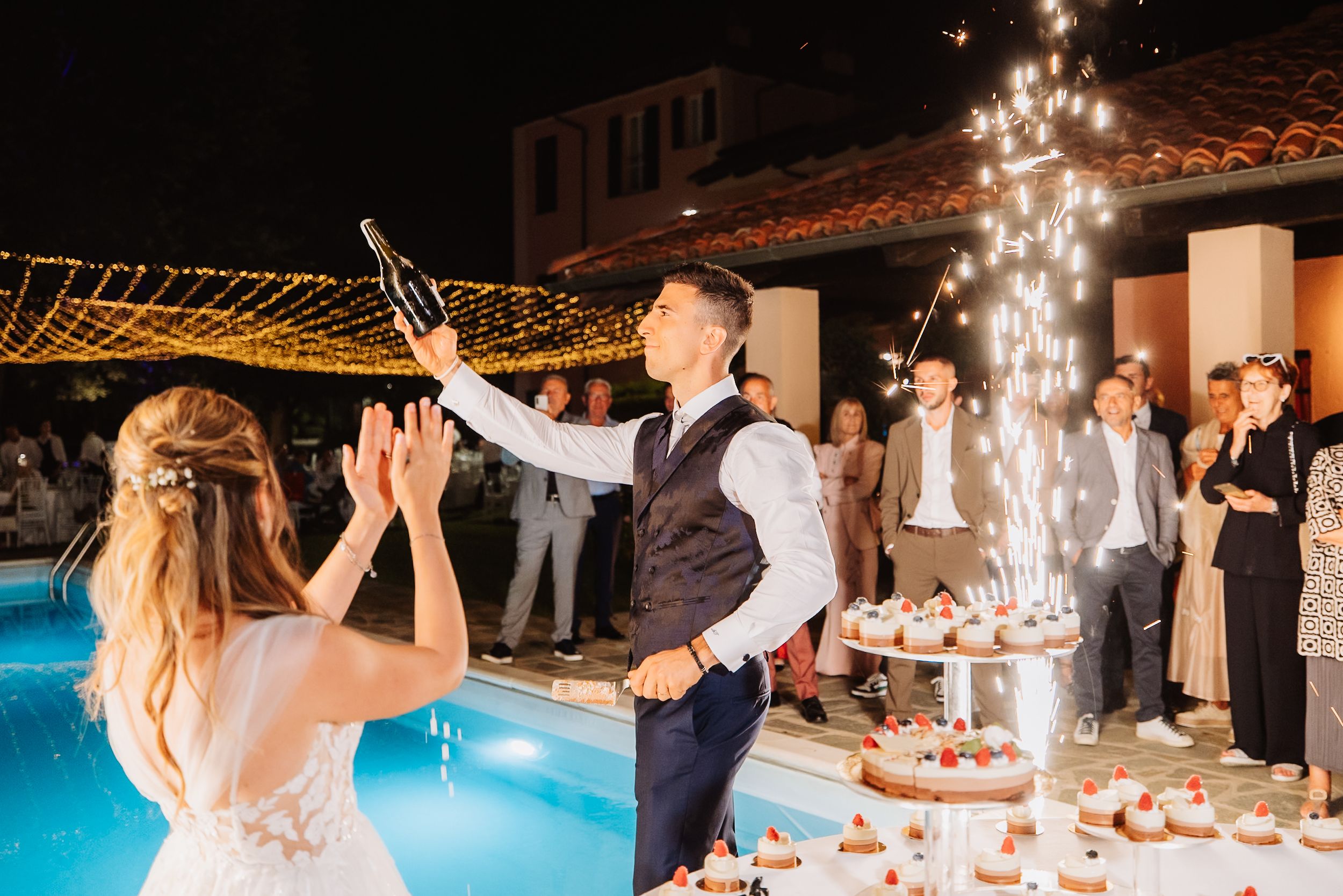 A romantic wedding reception celebration at night with sparklers, dancing and a poolside venue featuring twinkling lights.