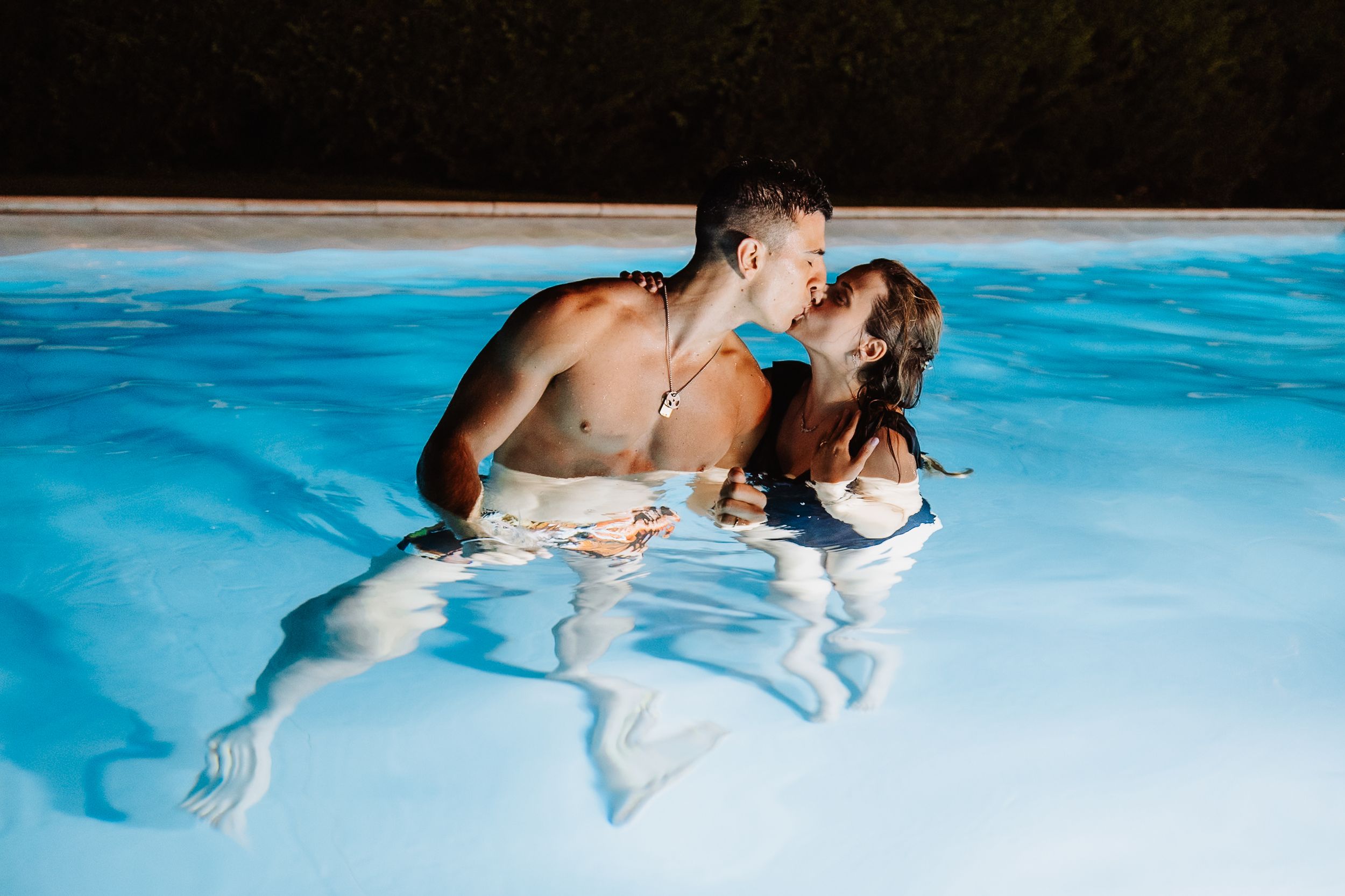 Couple swimming and embracing in a bright blue swimming pool with rippling reflections in the water.