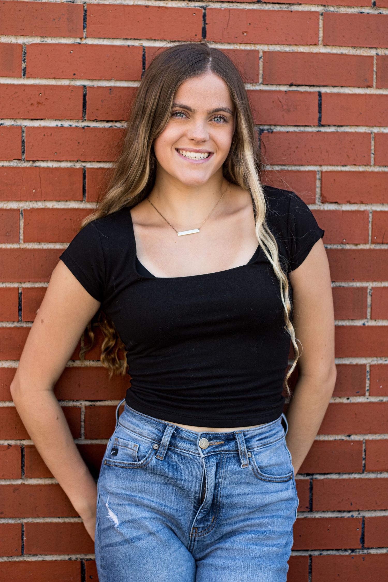 Person wearing black top and blue jeans posing casually against red brick wall outdoors.