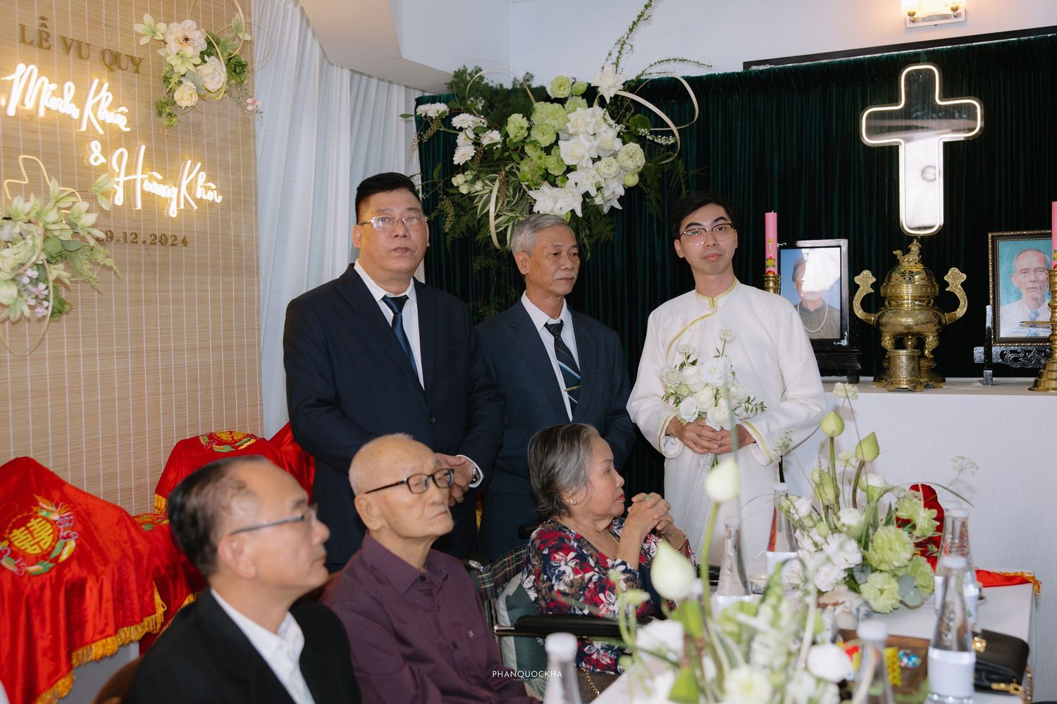 Vietnamese wedding guests and groom in white ao dai stand near altar with cross and floral arrangements during ceremony.