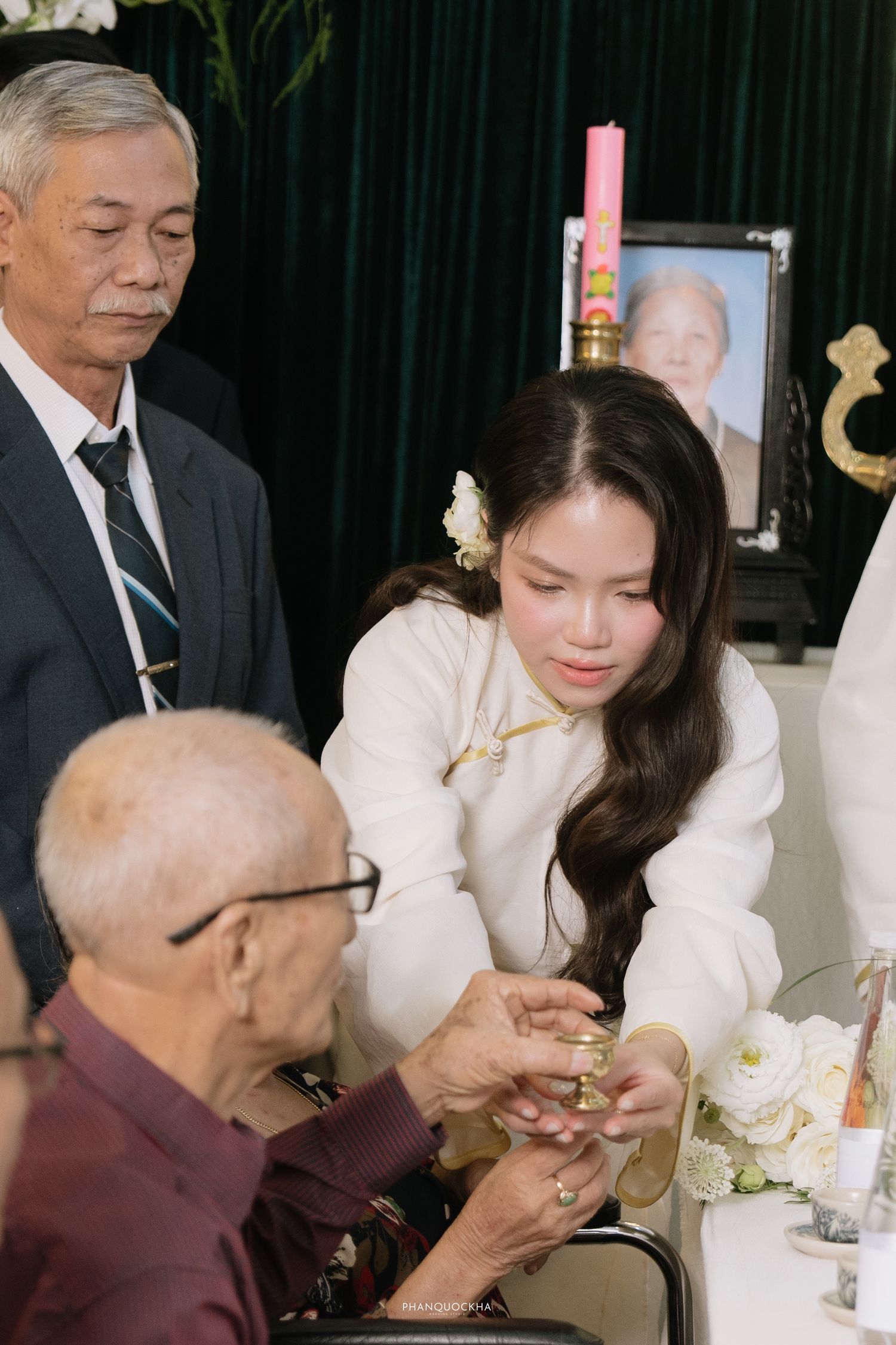 Young woman in white ao dai receiving jewelry from elderly relative at Vietnamese engagement ceremony.
