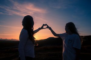 Two girls making a heart shape with their hands against a colorful sunset sky.