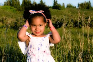 Cute young girl with afro puff hairstyle in white dress posing in green meadow.