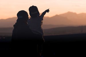 Silhouette of parent holding child pointing at mountain horizon during golden sunset.