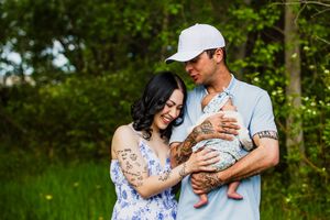 Young couple with tattoos holding newborn baby outdoors in green nature setting.
