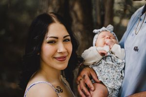 Young woman with dark hair smiling while holding newborn baby in floral bonnet outdoors.
