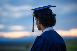Graduate in cap and gown gazing at a scenic sunset landscape from behind.