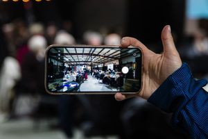 Person holding smartphone photographing crowded banquet hall event from audience perspective.