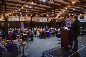 Speaker at podium addressing large seated audience in rustic barn venue with string lights.