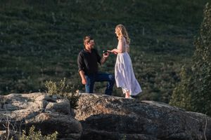 Man proposing to woman on rocky outcrop with scenic green hillside backdrop at sunset.