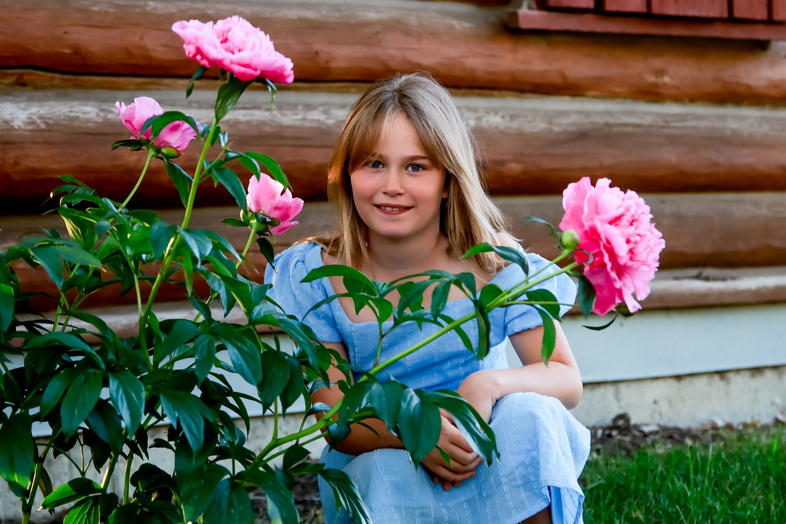 Young girl with blonde hair smiling beside blooming pink peonies near a log cabin outdoors.