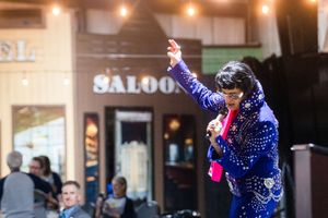 Elvis impersonator in blue sequin jumpsuit performing outside saloon with arm raised.