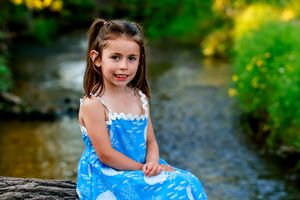 A young girl in a blue dress sits by a creek, smiling sweetly in a lush green outdoor setting.