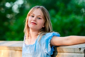 Young blonde girl in a blue dress posing outdoors with green trees behind her.
