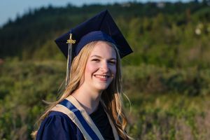Smiling young woman in navy blue graduation cap and gown posing outdoors with green hills in the background.