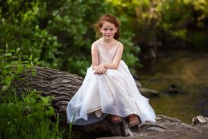 A young girl in a white ball gown sits gracefully on rocks near a stream surrounded by lush green foliage.
