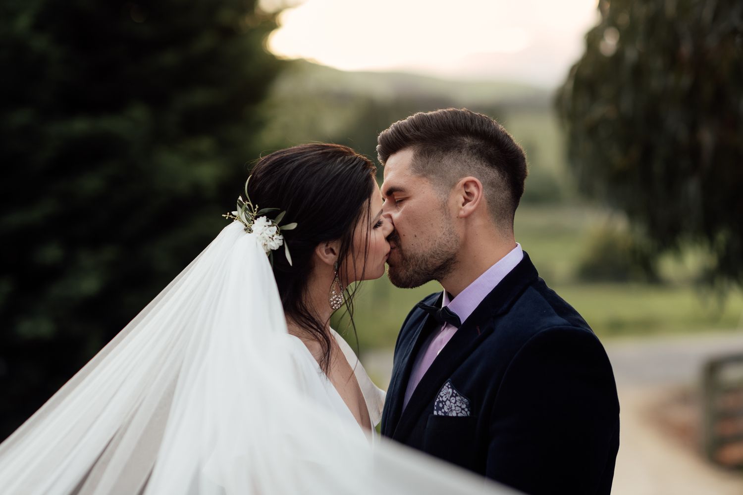A bride and groom kiss with a valley of wines in the background at their outdoor wedding in Martinborough by August & Vine