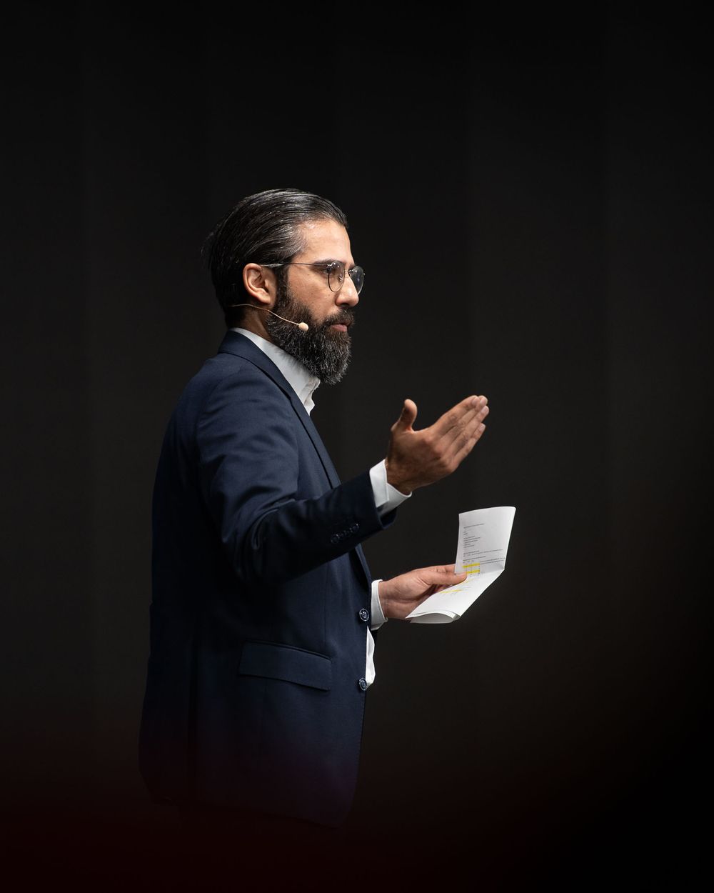 Man in a navy suit lit against dark background takes a sip from a small white paper cup.