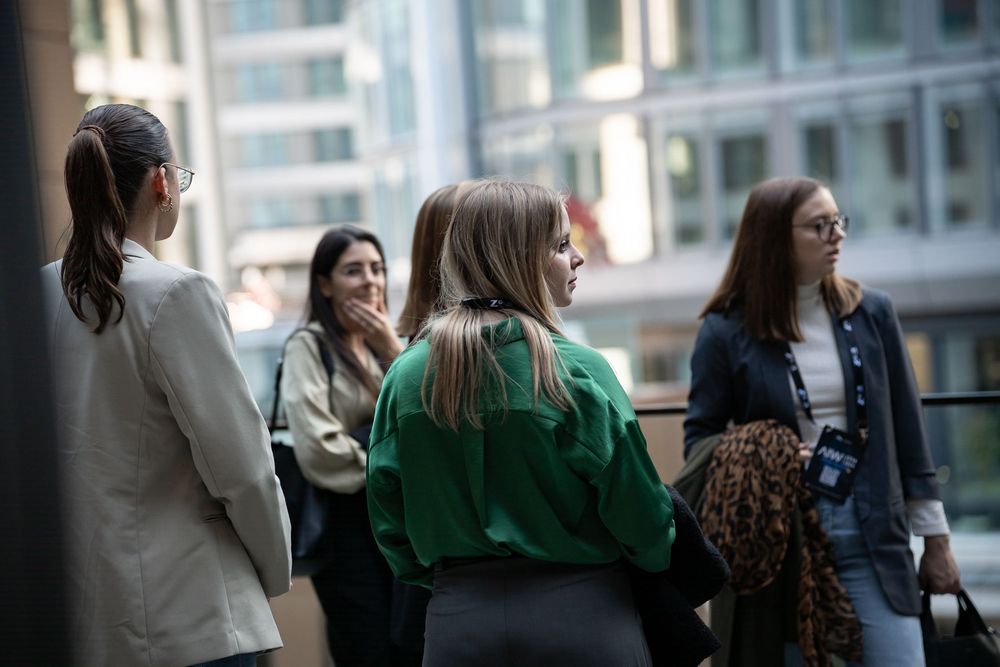A group of people gather near windows in an office or business setting having a casual conversation.
