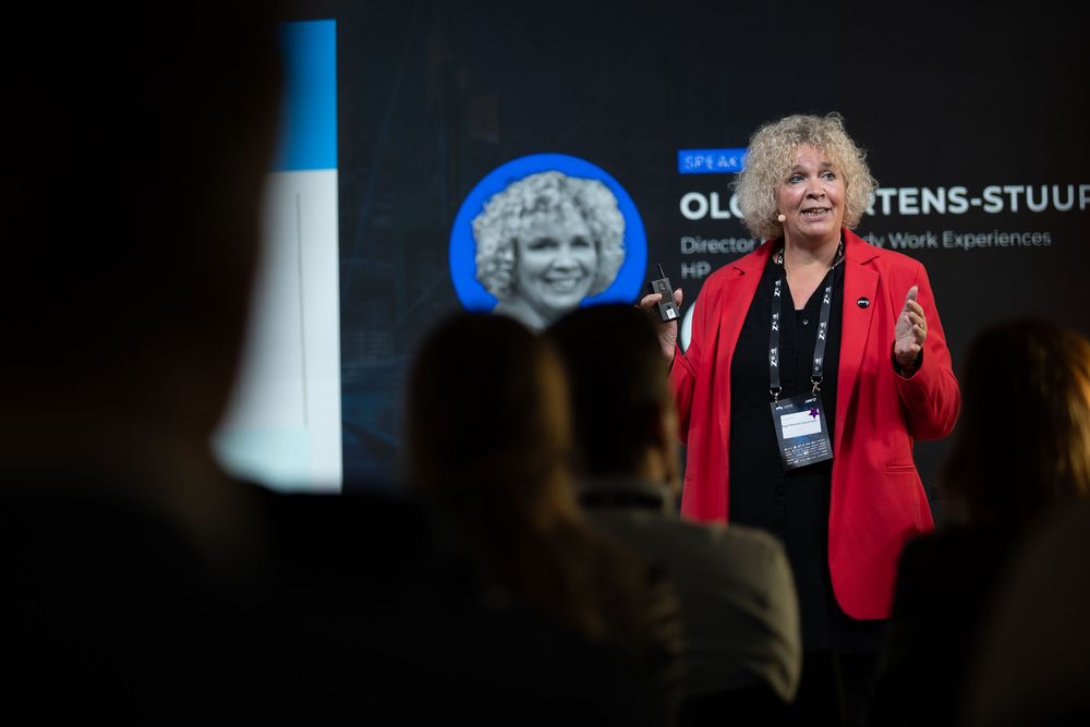 A person in a bright red jacket gestures while speaking at a podium during a presentation.