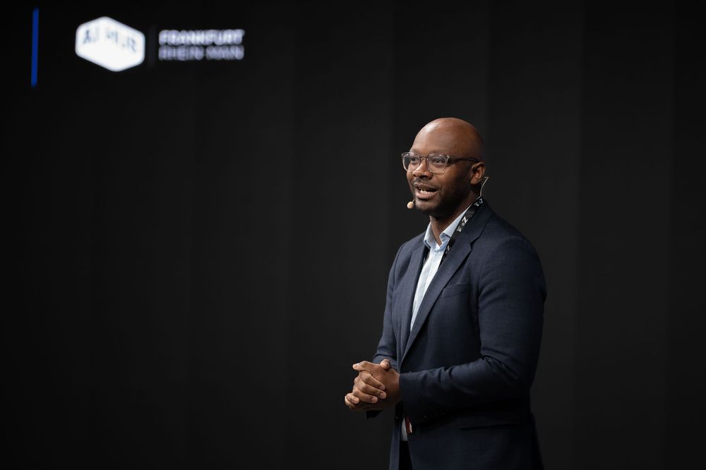 Professional speaker in business attire presenting on stage at a conference event with large display screens behind them.