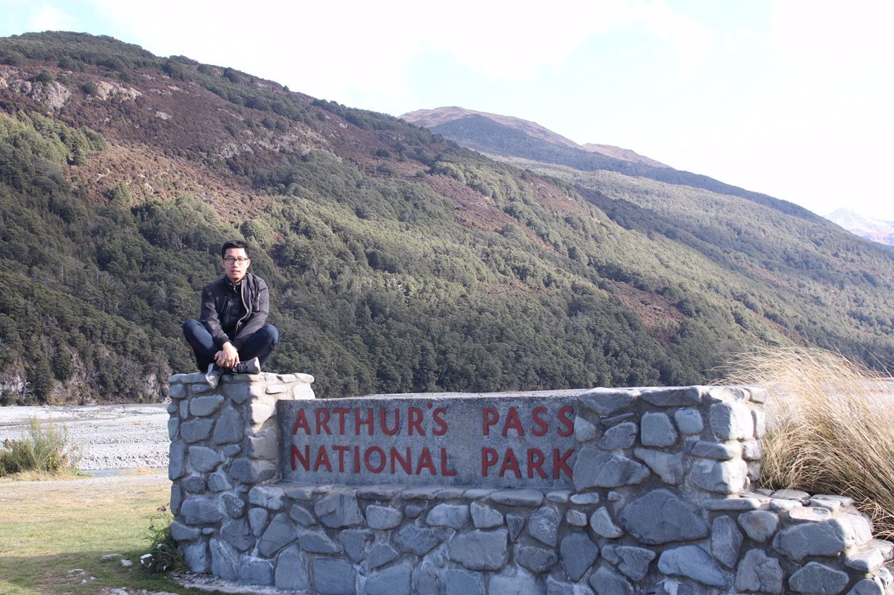 Stone wall entrance sign welcomes visitors to Arthurs Pass National Park against mountainous backdrop in New Zealand.
