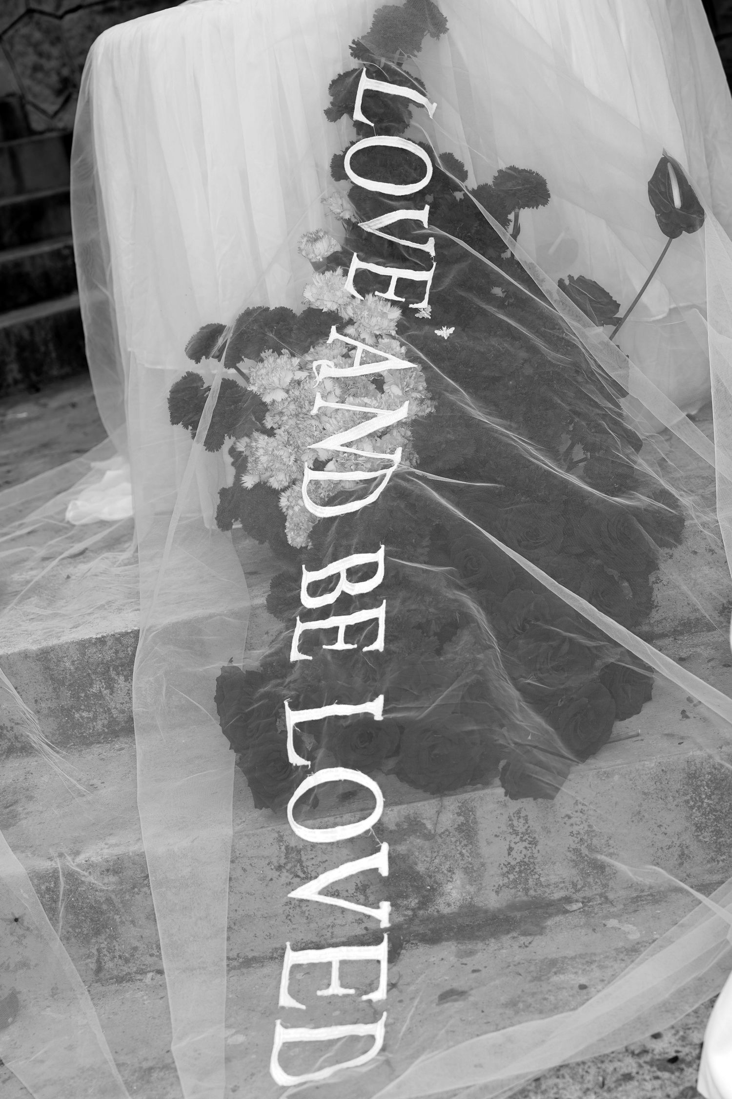 Black and white wedding veil with Love And Be Loved text draped over dark floral bouquet.