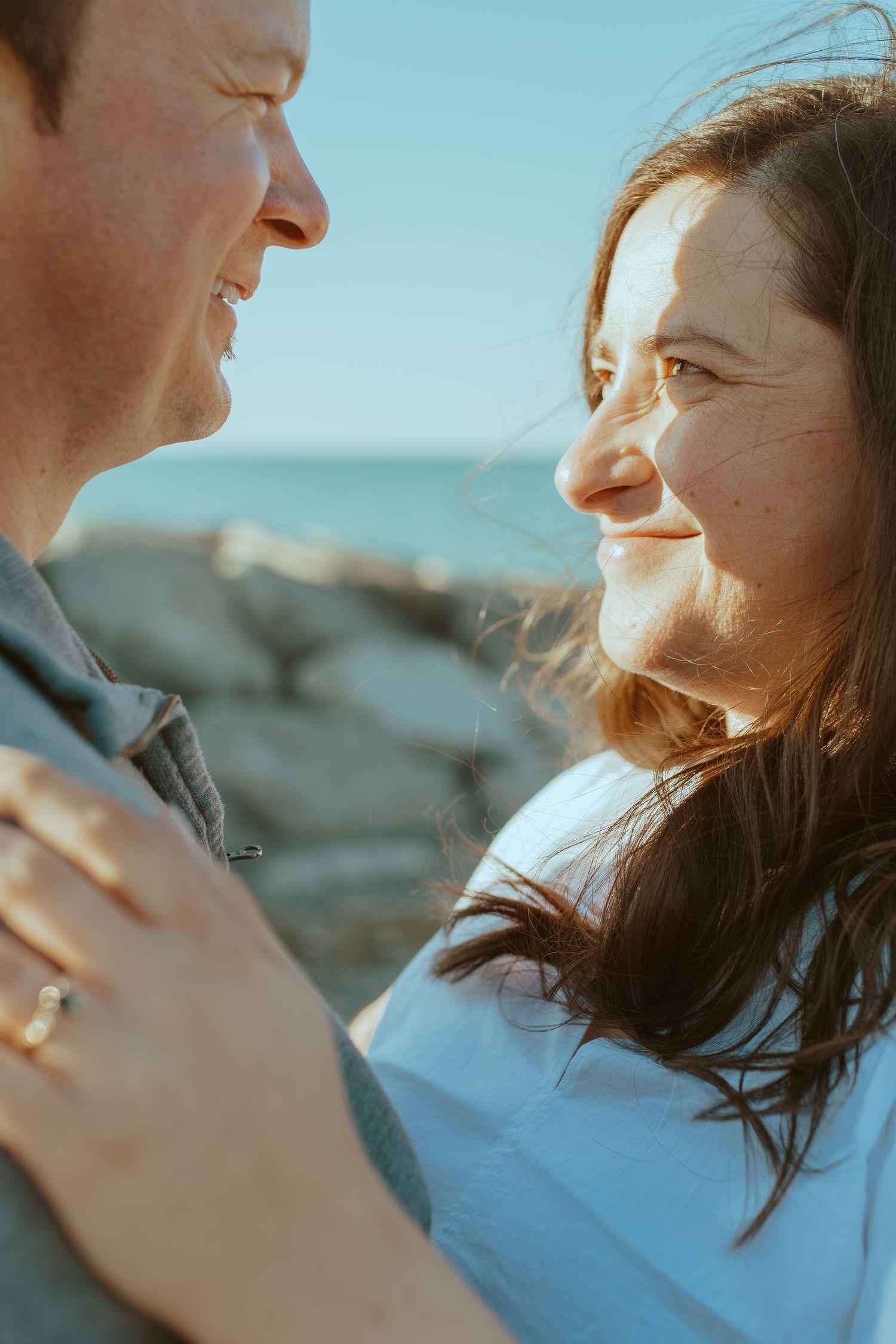 A loving couple shares an intimate moment smiling at each other against a blurred coastal background.