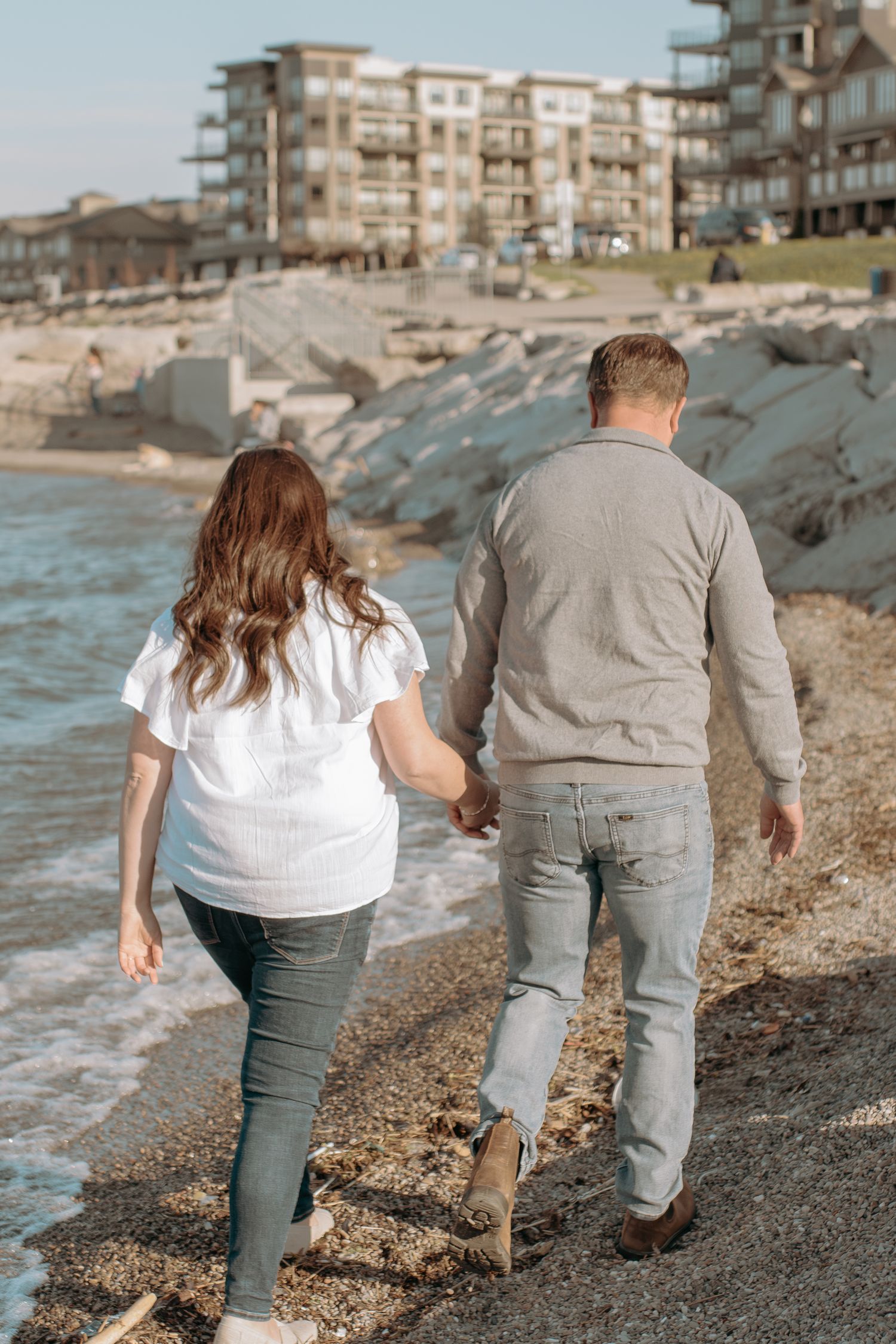 Two people walking together along a rocky beach shoreline with waterfront condominiums in the background.