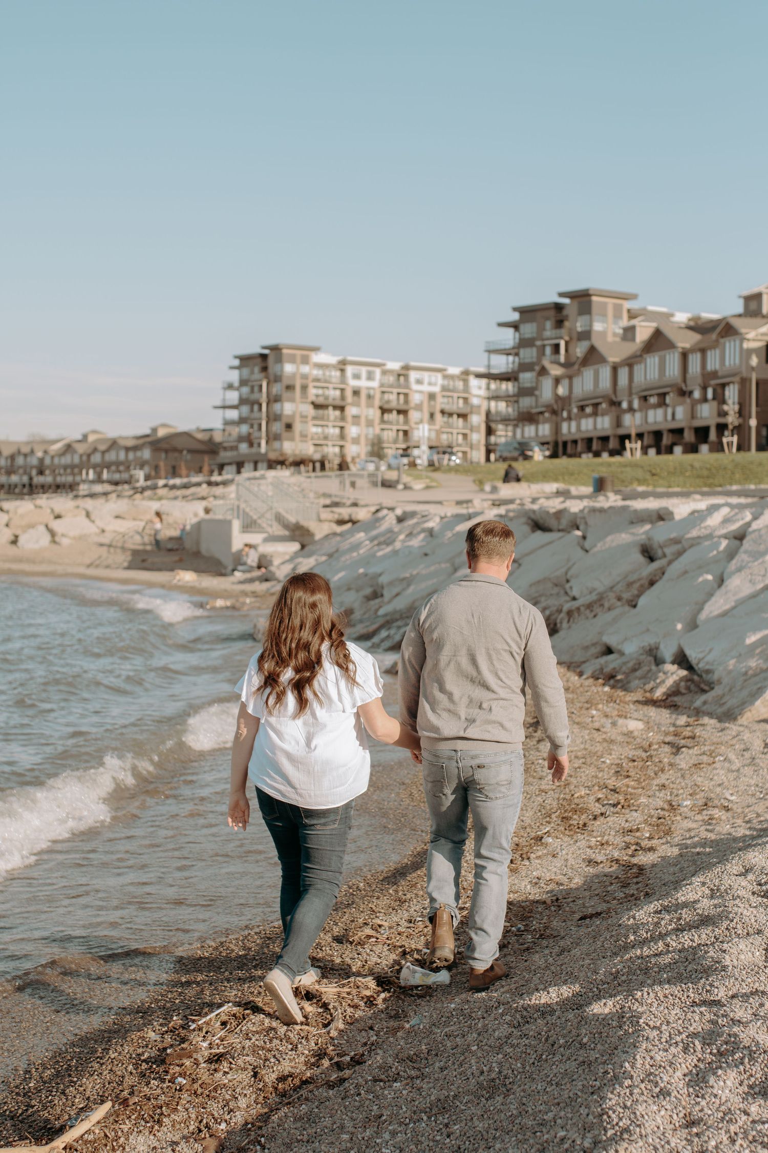 A couple walks hand in hand along a rocky beach with coastal buildings in the background.