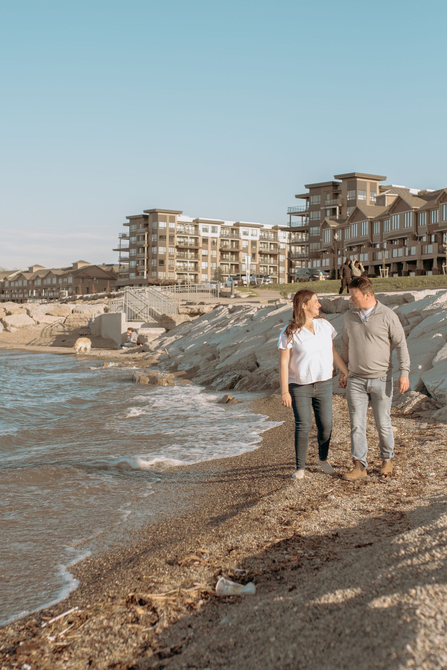 A couple strolls along a scenic beach with waves and coastal apartments in the background at sunset.