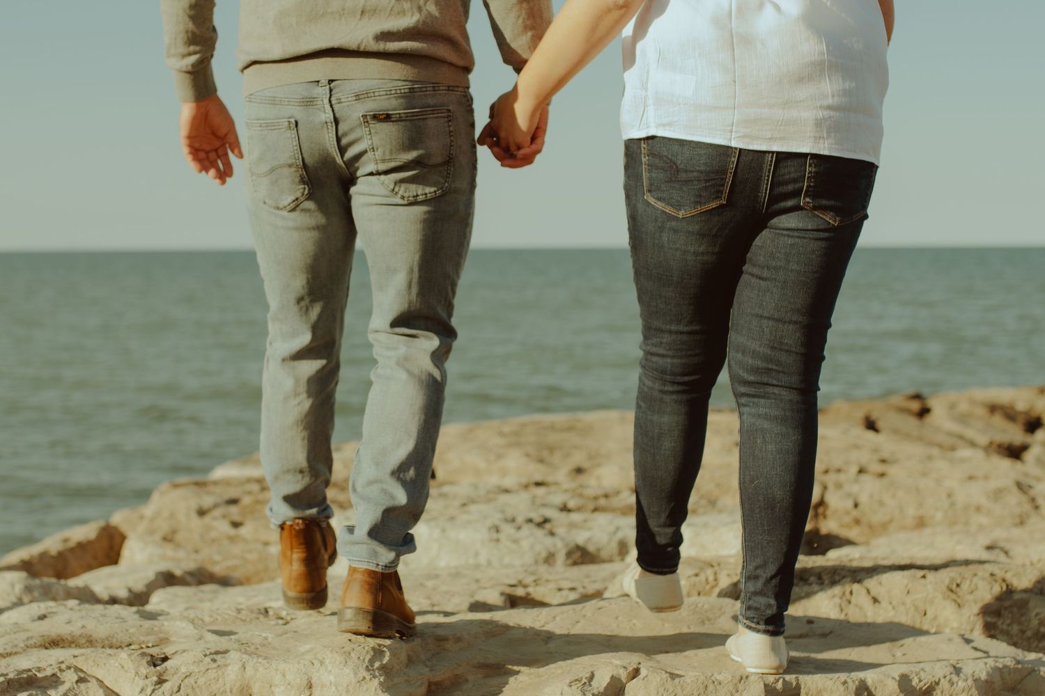 Two people walk barefoot along a sandy beach shoreline at sunset with ocean waves visible in the background.