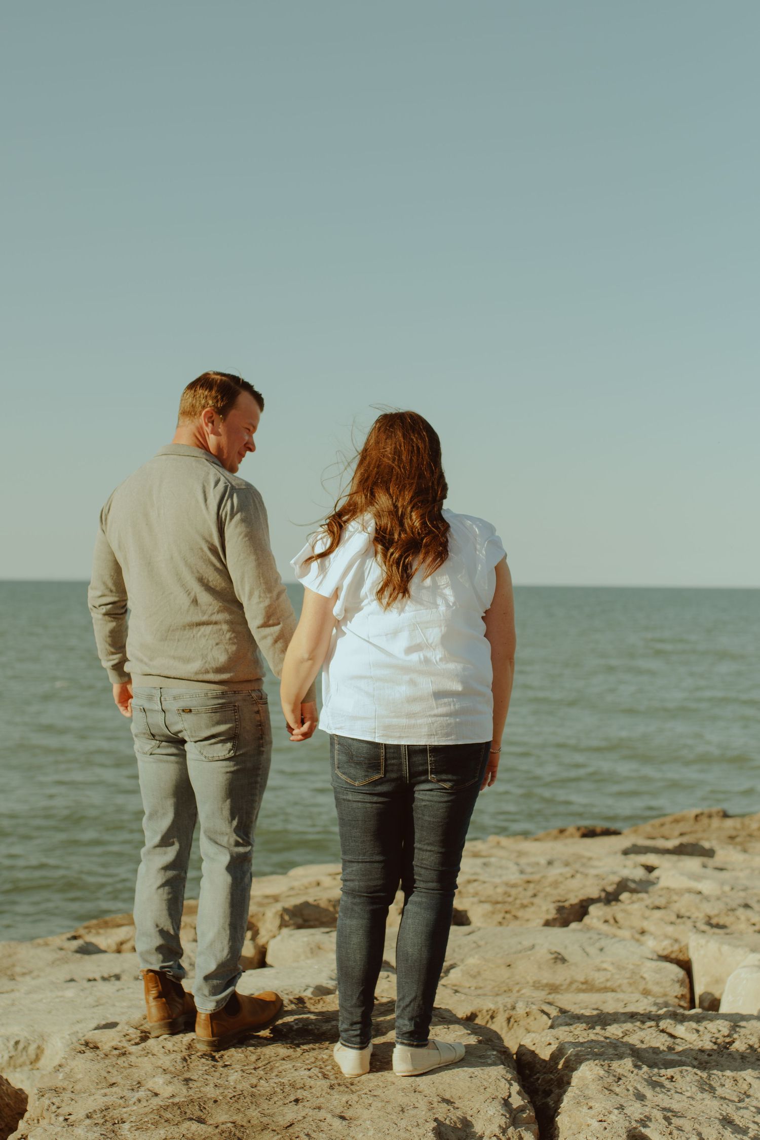 A couple holds hands while standing together on rocky coastal shoreline at sunset.