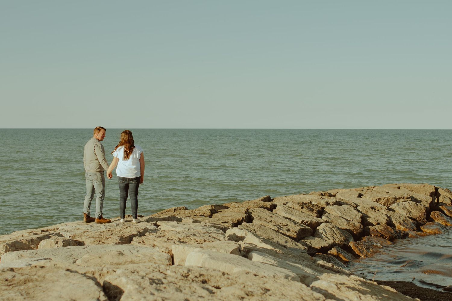 Two people walk along a rocky jetty extending into the tranquil ocean on a sunny afternoon.