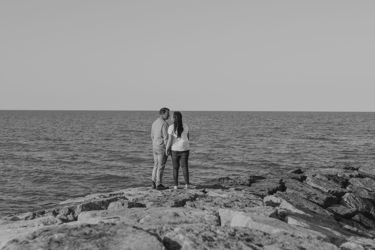 A couple stands on rocky coastal outcrop overlooking ocean in black and white photography.
